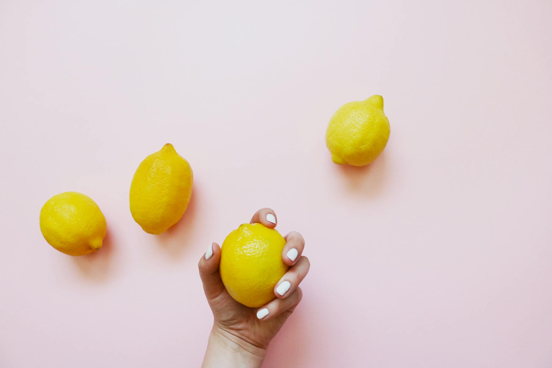 Eine Hand mit weißen Nägeln, die eine Zitrone auf zartem rosa Hintergrund hält, umgeben von drei weiteren Zitronen