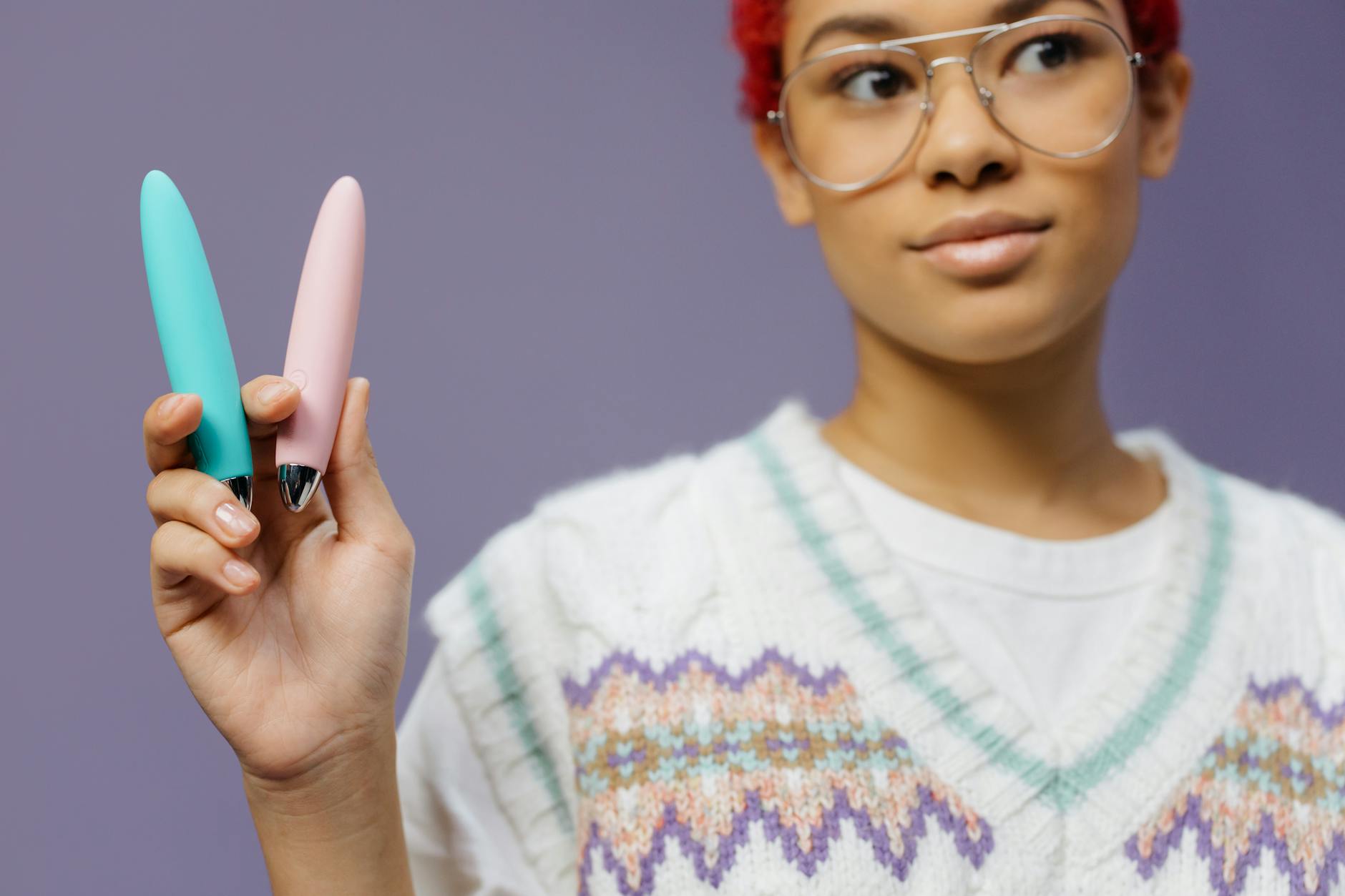 Eine junge Frau mit Brille hält zwei bunte Vibratoren gegen einen lila Hintergrund.